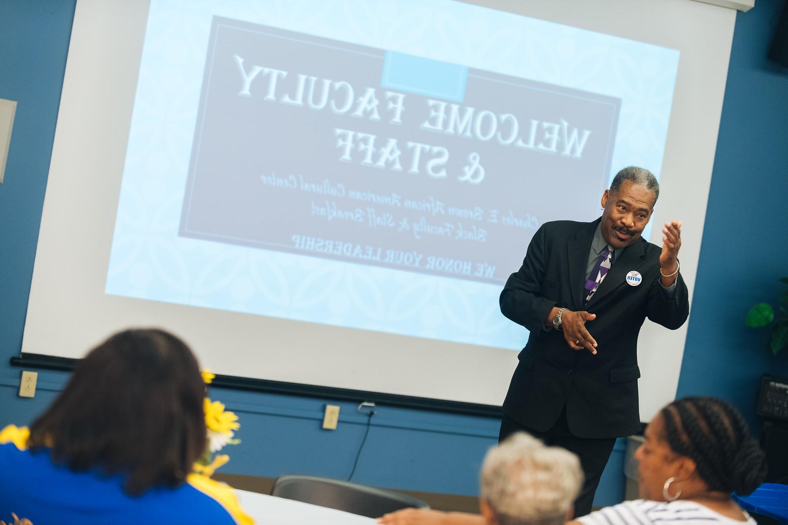 A man in a suit speaks in front of a group of faculty and staff with a presentation slide in the background that says 'Welcome Faculty & Staff